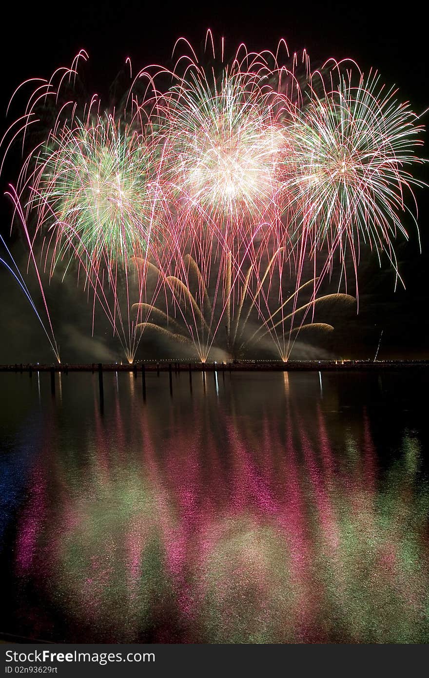 Long exposure photo of multiple fireworks over a lake, against a black sky.