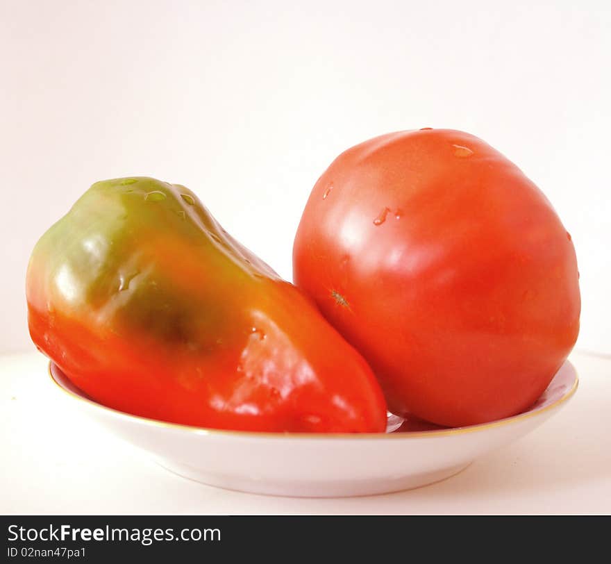 Red pepper and tomato on a saucer,one large red pepper next to a big tomato on a saucer and vegetables on the plate. Red pepper and tomato on a saucer,one large red pepper next to a big tomato on a saucer and vegetables on the plate