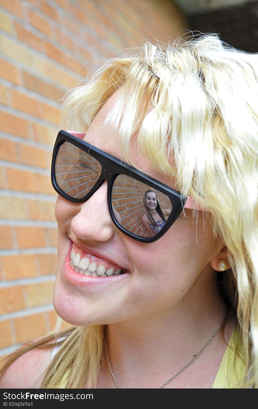 A girl smiling can be seen in her friend's sunglasses reflection against a brick wall. A girl smiling can be seen in her friend's sunglasses reflection against a brick wall.