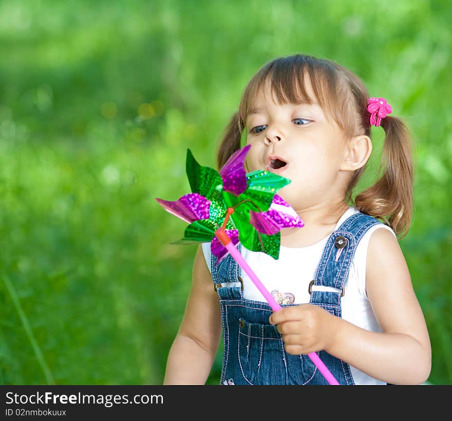 Little girl in jeans blowing on color propeller outdoor on green background. Little girl in jeans blowing on color propeller outdoor on green background