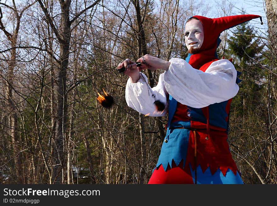 Artistic man in a suit of fire-clown rotate a fiery chain posing on backround of forest. Photo.