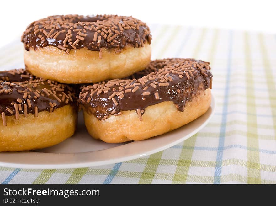 Three chocolate donuts with glazing and sprinkles on top, placed on round plate and linen cloth in green and blue stripes. Three chocolate donuts with glazing and sprinkles on top, placed on round plate and linen cloth in green and blue stripes