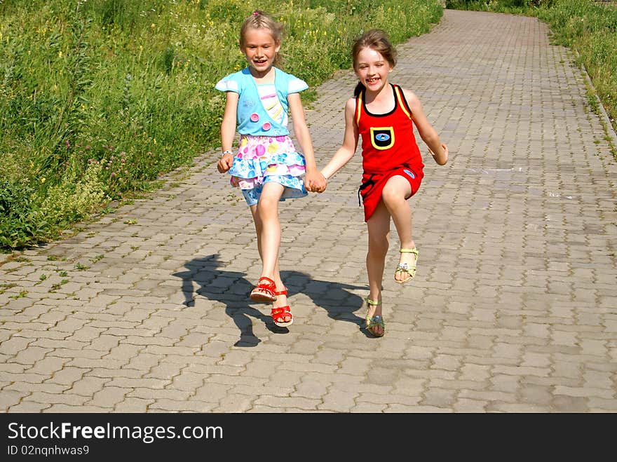 Two joyful girls keep for hands and run on sidewalk jumping up in the summer. Two joyful girls keep for hands and run on sidewalk jumping up in the summer