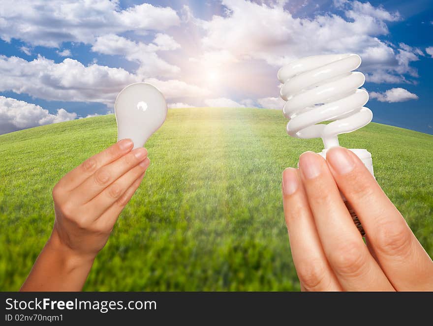 Female Hands Holding Energy Saving and Regular Light Bulbs Over Arched Horizon of Grass Field, Clouds and Sky. Female Hands Holding Energy Saving and Regular Light Bulbs Over Arched Horizon of Grass Field, Clouds and Sky.