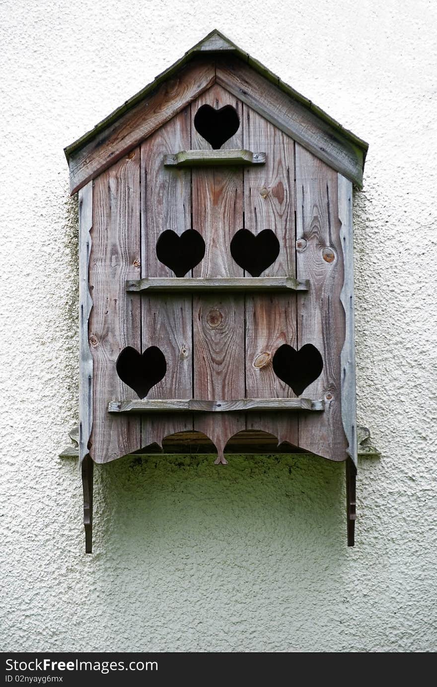 A vertical image of an ornate birdbox with heart shaped openings on a white wall. A vertical image of an ornate birdbox with heart shaped openings on a white wall