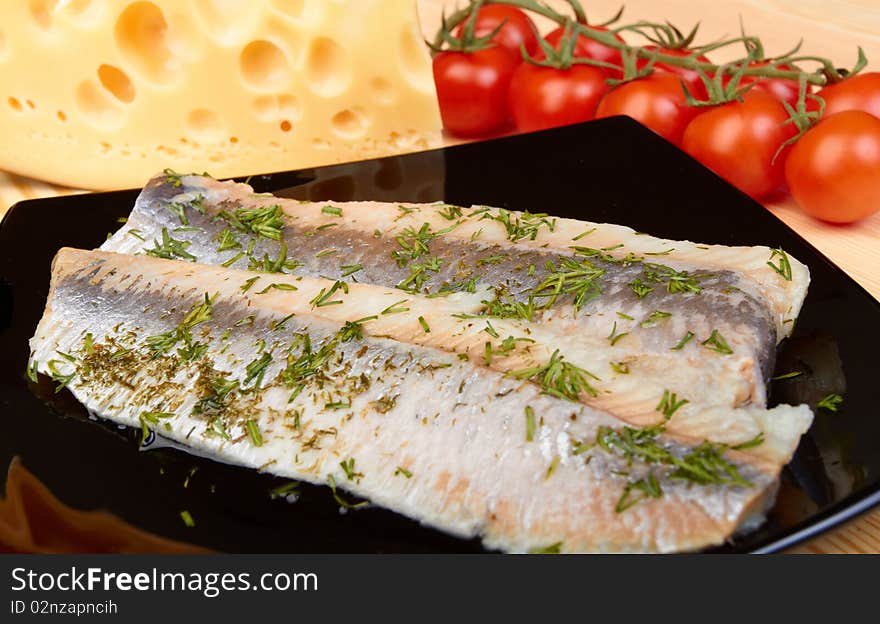 Herring on black plate with piece of cheese and tomato cherry on wood board