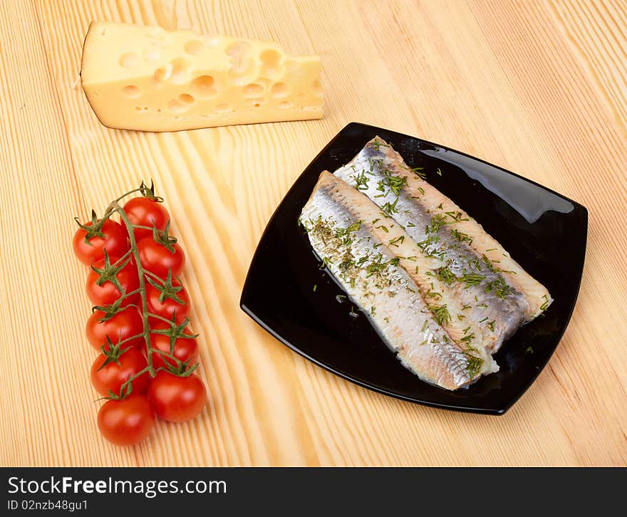 Herring on black plate with piece of cheese and tomato cherry on wood board