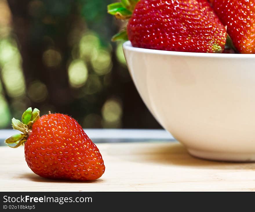 A single strawberry next to a white bowl of strawberries on a cutting board. A single strawberry next to a white bowl of strawberries on a cutting board