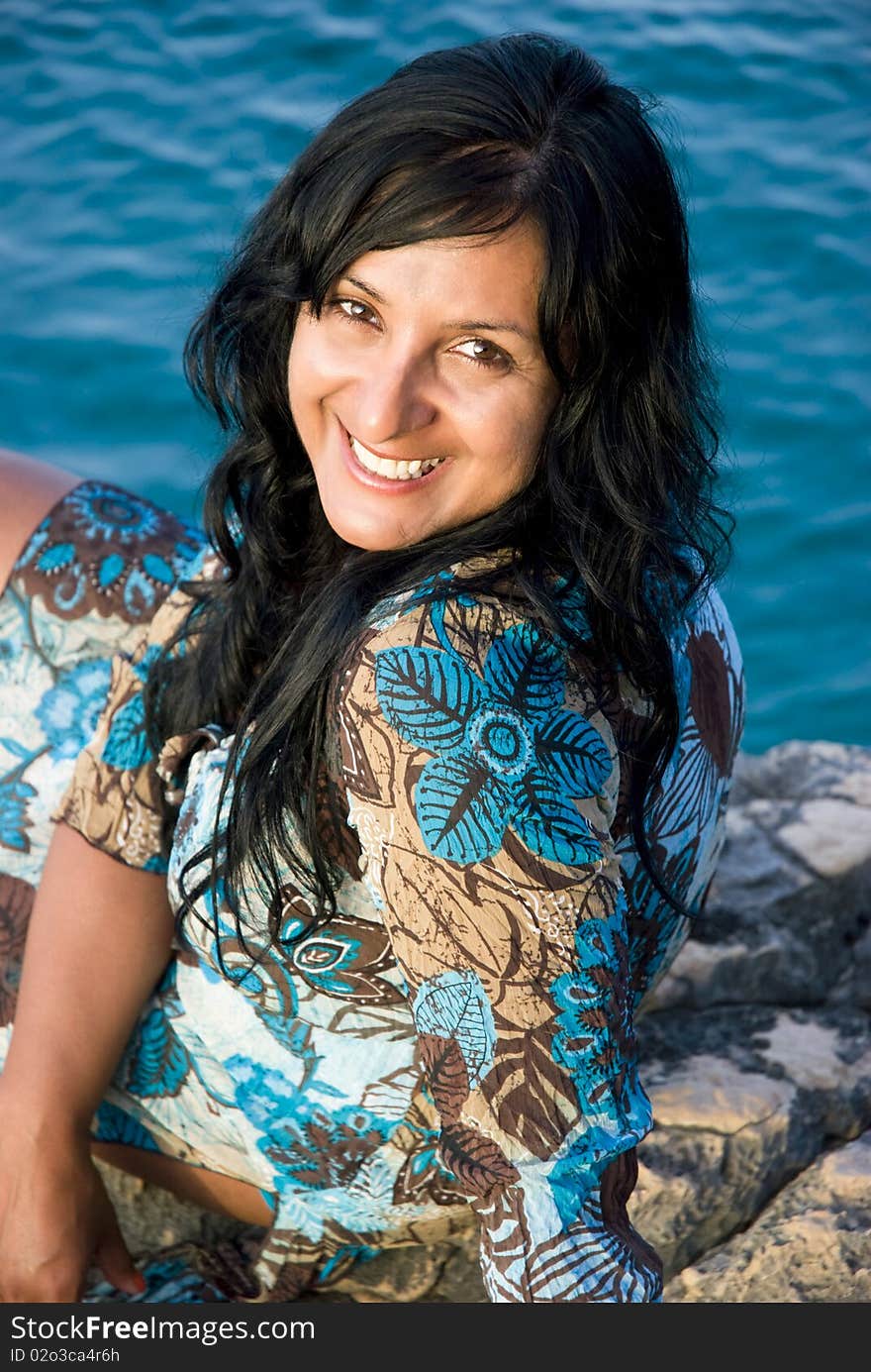 A beautiful happy smiling indian woman sitting on a rocky beach in Paxos Greece.