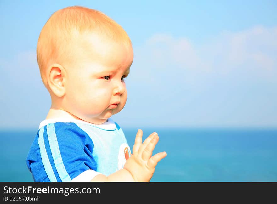 Beautiful white little boy in a bright blue t-shirt is seriously looking into the future put out his hand against the background of summer sea and blue sky. Beautiful white little boy in a bright blue t-shirt is seriously looking into the future put out his hand against the background of summer sea and blue sky