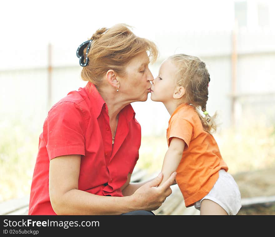 Grandma kisses granddaughter. They sit under the open sky on a summer day. Grandma kisses granddaughter. They sit under the open sky on a summer day