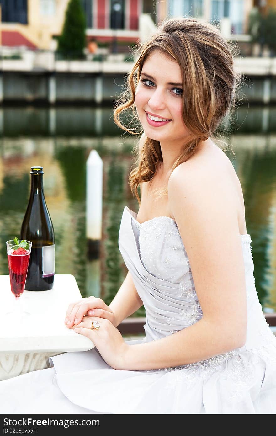 An attractive young woman is smiling and wearing formal attire while sitting at a dockside table with an alcoholic beverage. Vertical shot. An attractive young woman is smiling and wearing formal attire while sitting at a dockside table with an alcoholic beverage. Vertical shot.