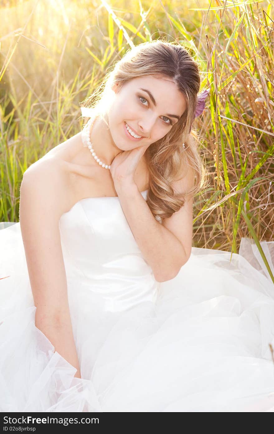 An attractive young bride wearing a white wedding dress and pearls is sitting with her head on her hand in the grass. Vertical shot. An attractive young bride wearing a white wedding dress and pearls is sitting with her head on her hand in the grass. Vertical shot.