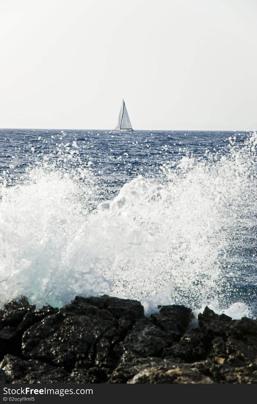 A wave hits the rocky shore and a sailing boat moves along in the backround in Paxos Greece. A wave hits the rocky shore and a sailing boat moves along in the backround in Paxos Greece.