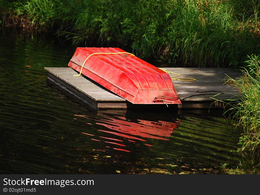 A red small row boat out of water upside down on a dock in a pond. A red small row boat out of water upside down on a dock in a pond.