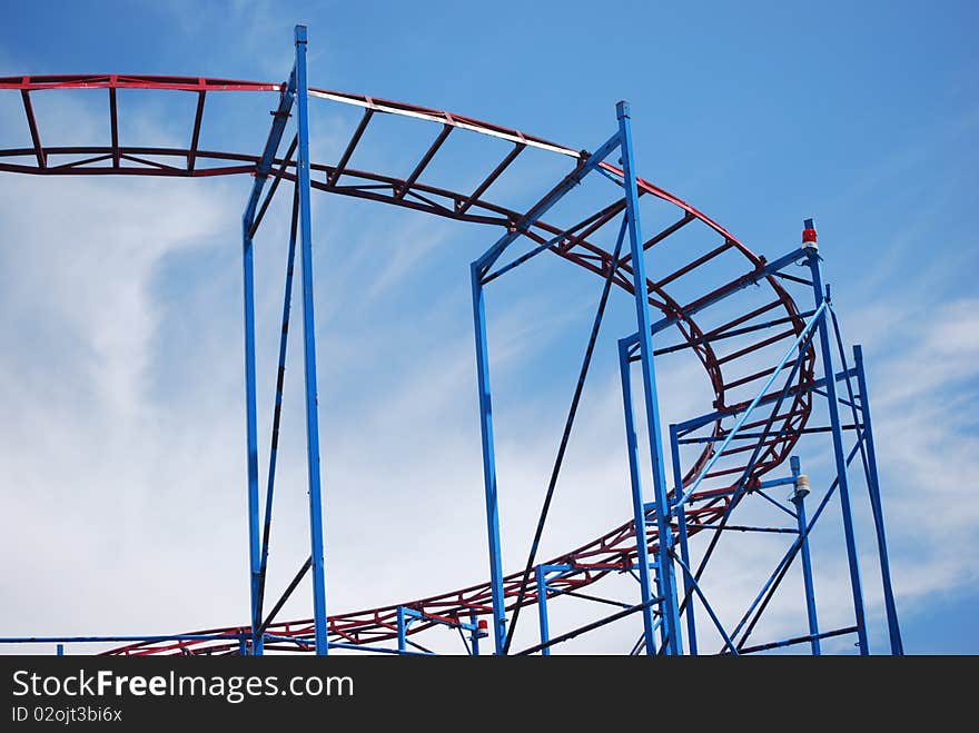 The curve of older metal roller coaster tracks that are red and blue taken at the Sylvan Beach New York amusement park on Lake Oneida with a cloudy blue sky as the background.