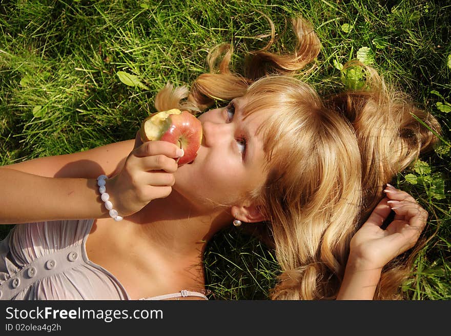 A beautiful young blonde with an apple lies on a grass in a city park