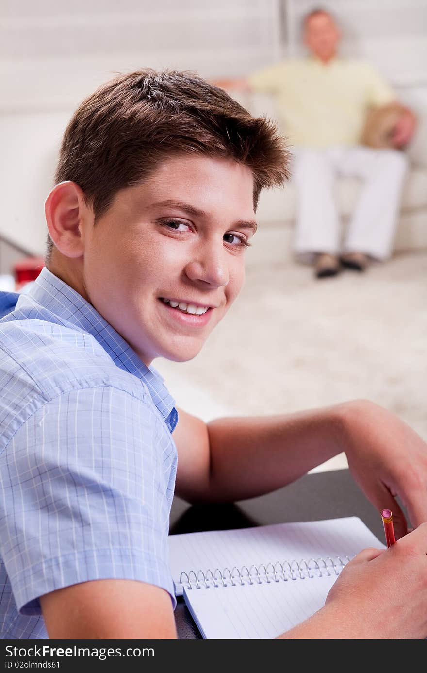 Close-up of young boy studying and looking at camera as father relaxes on the couch in the background. Close-up of young boy studying and looking at camera as father relaxes on the couch in the background