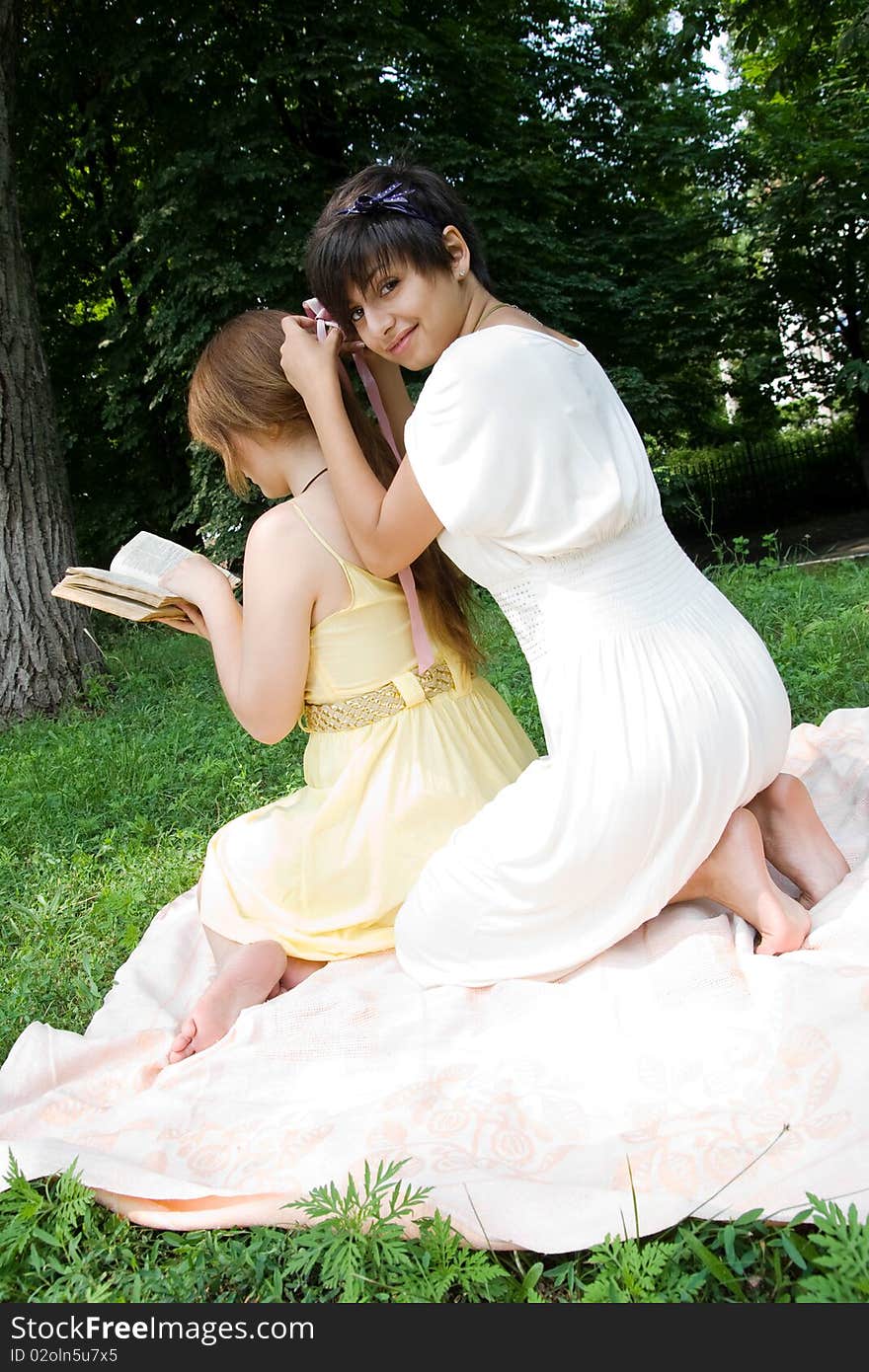 Two girls spending their free time outdoors. One is reading a book and the other is braiding her hair. Two girls spending their free time outdoors. One is reading a book and the other is braiding her hair.