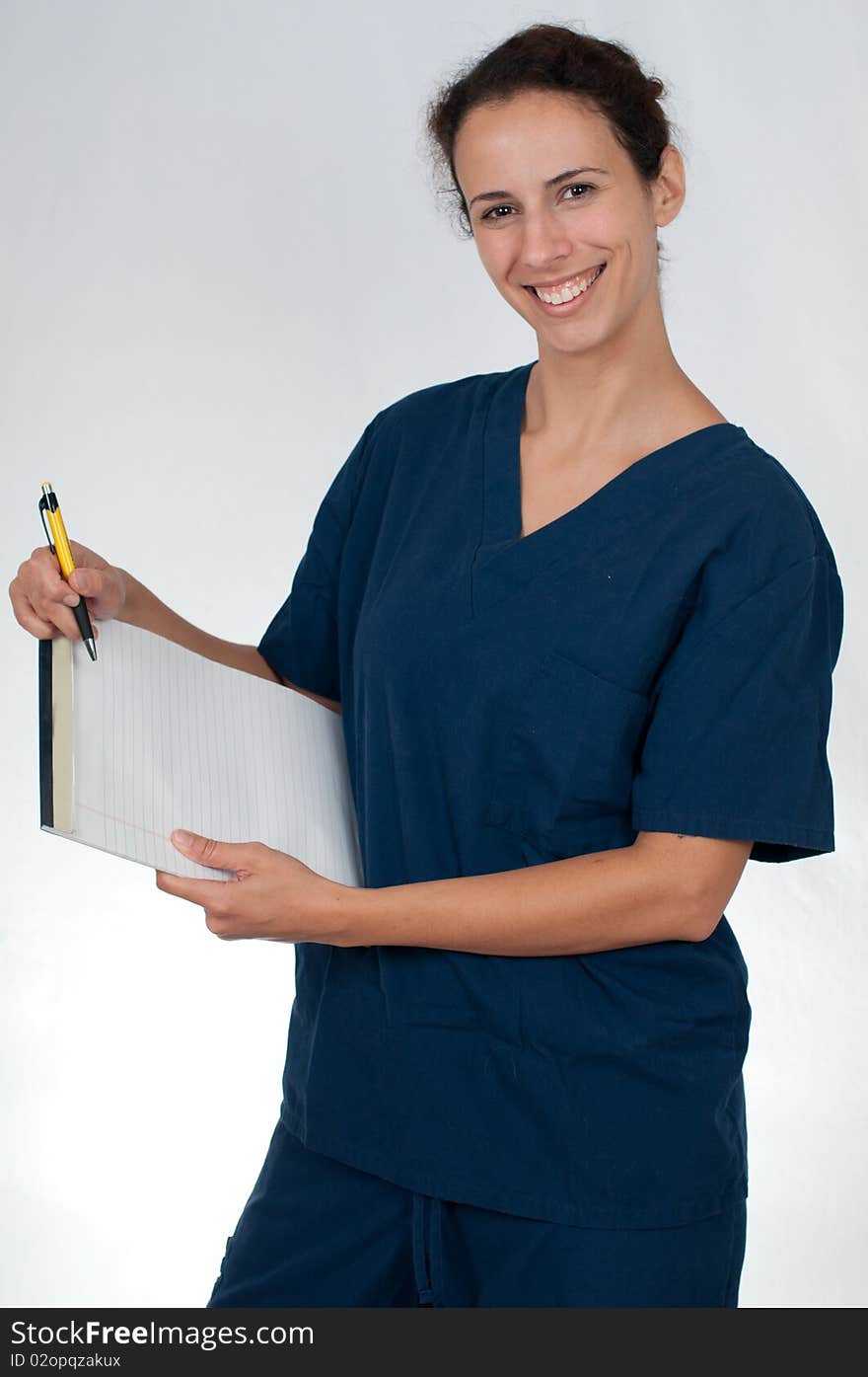 Brunette woman wearing blue scrubs against white background, holding tablet of paper and yellow pen. Brunette woman wearing blue scrubs against white background, holding tablet of paper and yellow pen