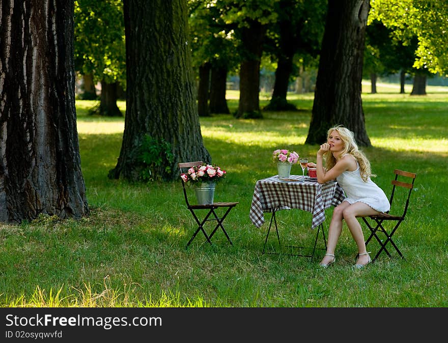 Young blond in the parc under trees, sitting relaxed on a folding chair beside a folding table with flowers and a wine glass on it. Young blond in the parc under trees, sitting relaxed on a folding chair beside a folding table with flowers and a wine glass on it