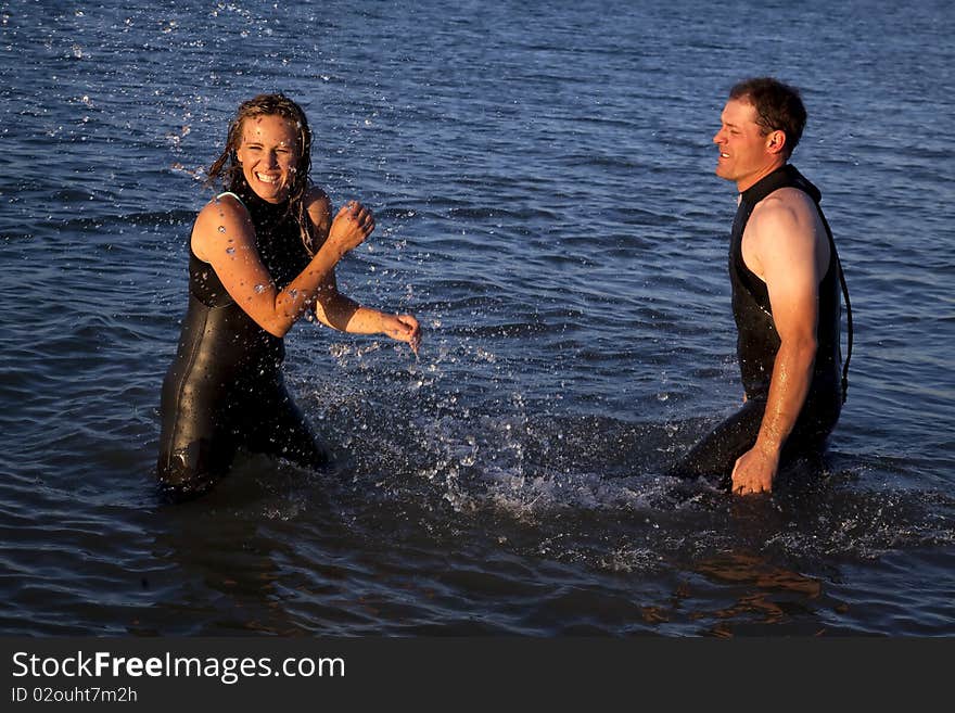 A couple in their wet suits playing and splashing around while they are standing in the water. A couple in their wet suits playing and splashing around while they are standing in the water.