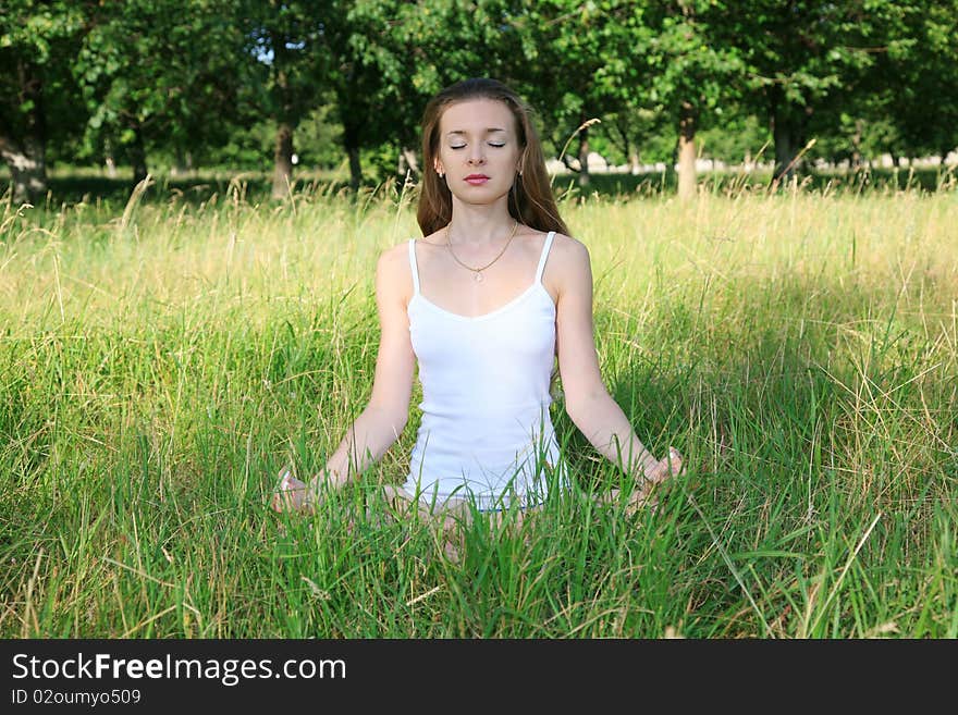 Beautiful young girl in white, sitting on the grass and meditate on the background of green trees. Beautiful young girl in white, sitting on the grass and meditate on the background of green trees