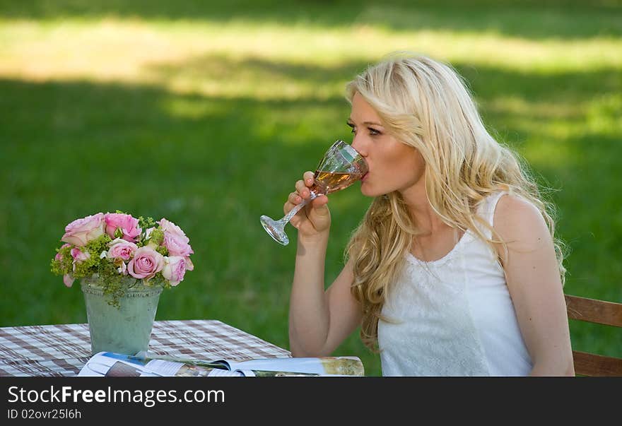 Young attractive blond with a glass of wine in her hand sitting in the green park on a folding table. Young attractive blond with a glass of wine in her hand sitting in the green park on a folding table