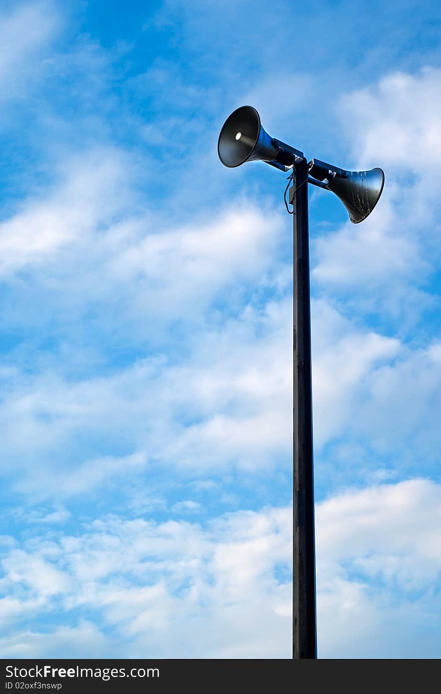 A public address system speakers tower against a cloudy, blue, daytime sky. A public address system speakers tower against a cloudy, blue, daytime sky.