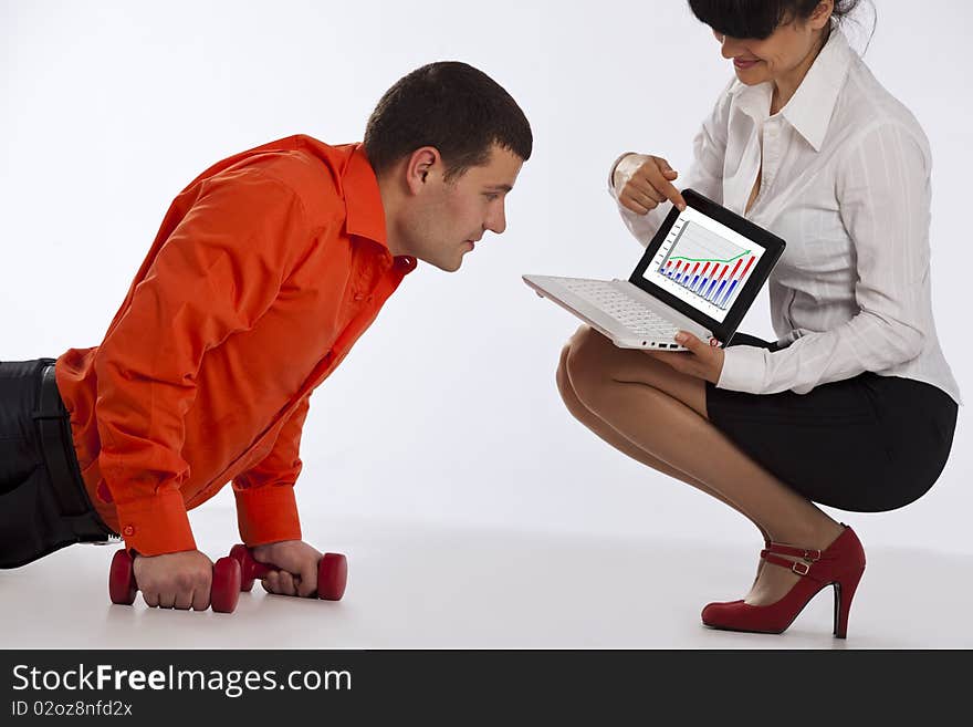 Young businessman doing push-ups with dumbbells, looking on laptop screen. His secretary, pointing to a successful business at the screen