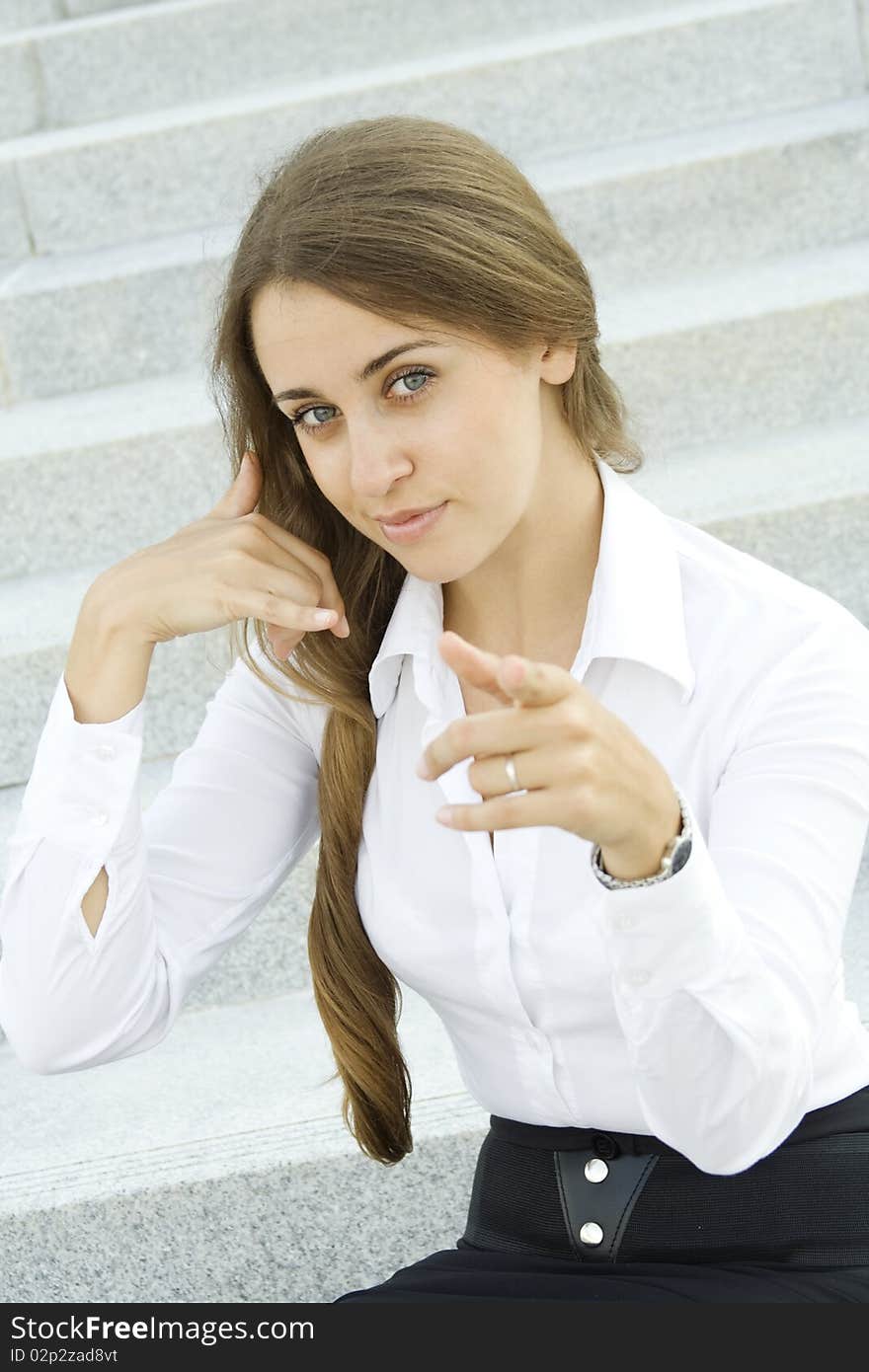 Young businesswoman outdoors on the stairs near the office building shows a sign of Call me pointing to viewer. Young businesswoman outdoors on the stairs near the office building shows a sign of Call me pointing to viewer