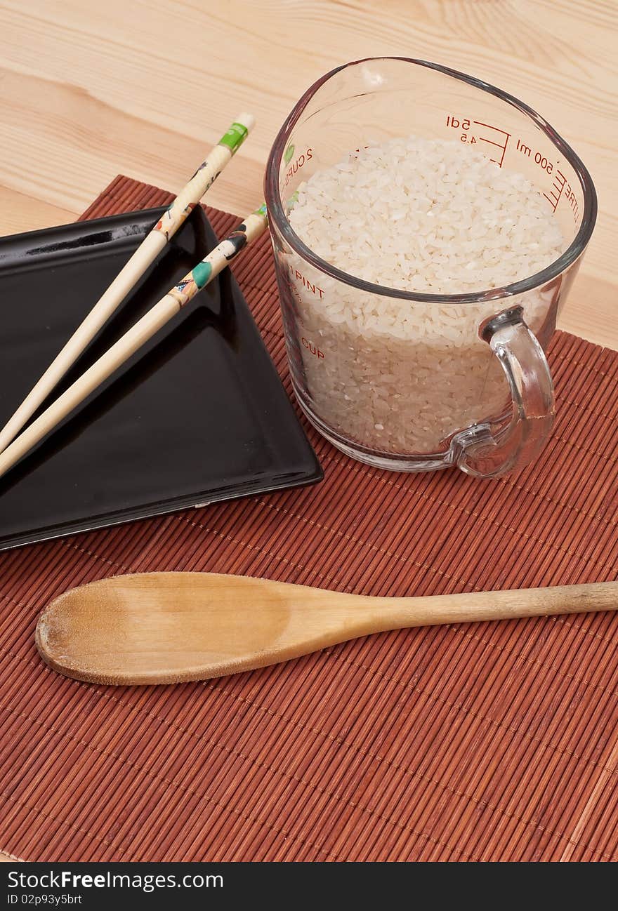 Cup Of Dry White Rice Next To Black Dish With Chop Sticks. Cup Of Dry White Rice Next To Black Dish With Chop Sticks