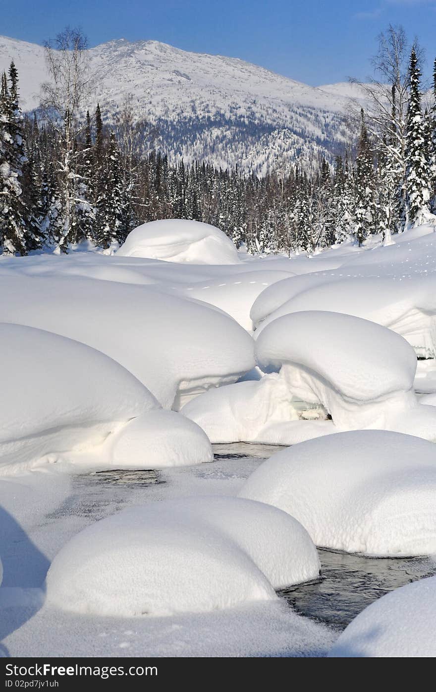 Thick fluffy snowdrifts on the banks and bed of mountain river. Pine forest and further mountain slope on background. Thick fluffy snowdrifts on the banks and bed of mountain river. Pine forest and further mountain slope on background.