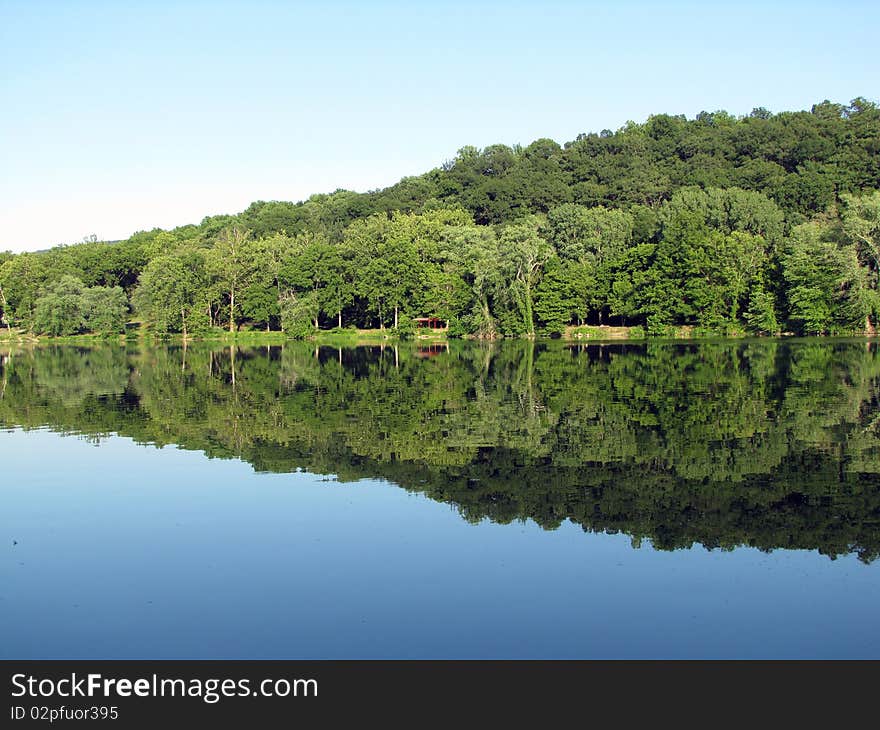 Green trees and a little red structure in the woods reflect vibrantly on a calm river. Green trees and a little red structure in the woods reflect vibrantly on a calm river.