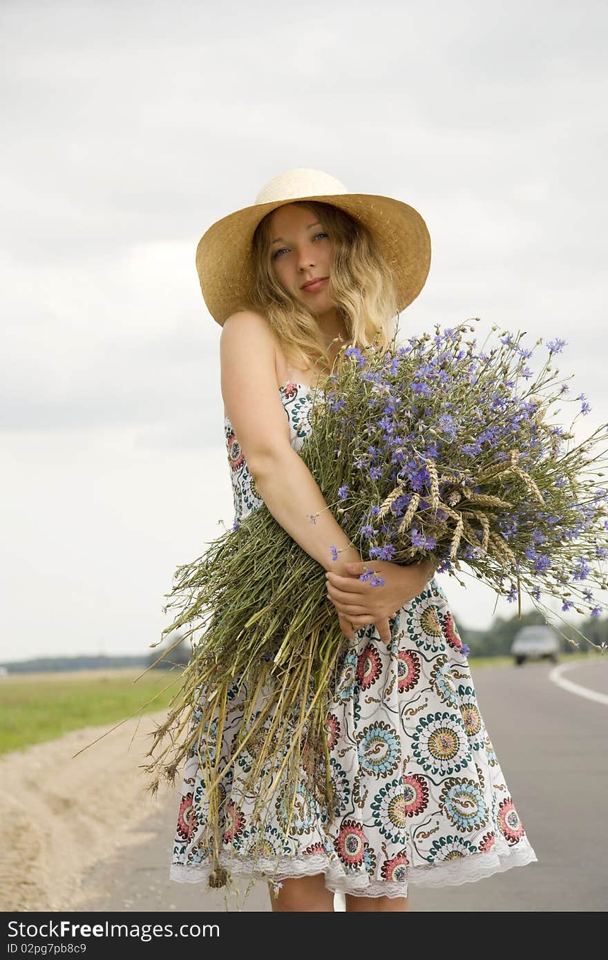 The woman in a hat with a bouquet of cornflowers costs on the brink of road near passing by the car. The woman in a hat with a bouquet of cornflowers costs on the brink of road near passing by the car