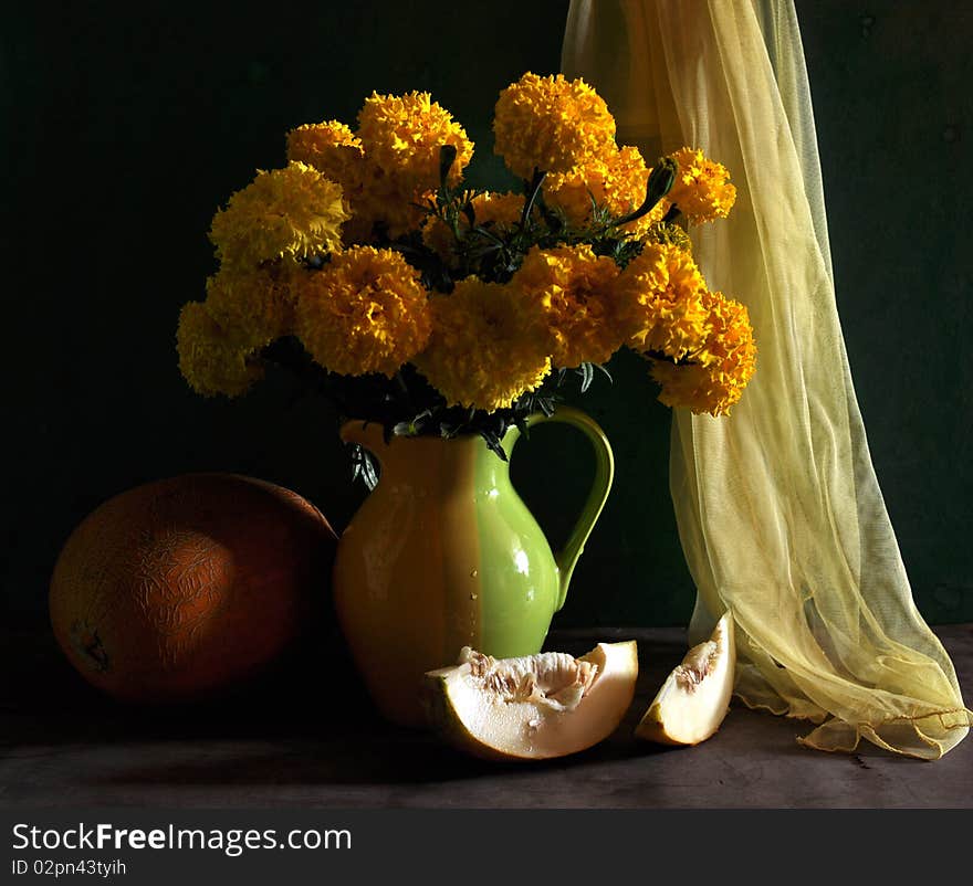 yellow marigolds are in a jug, a melon and shawl lie alongside. yellow marigolds are in a jug, a melon and shawl lie alongside