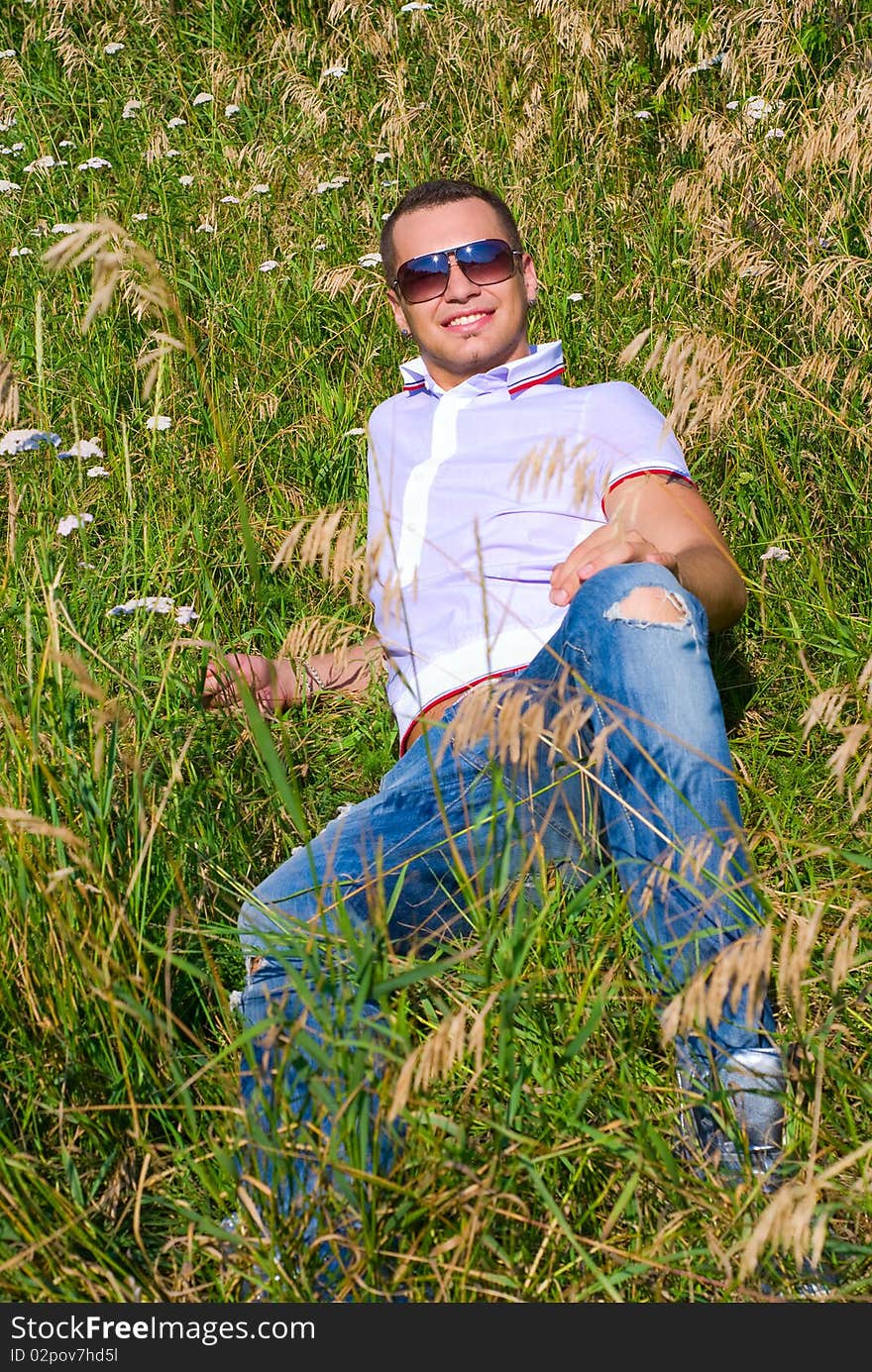 Young man in jeans with teeth smile is laying on summer field. Green grass surrounds him. Young man in jeans with teeth smile is laying on summer field. Green grass surrounds him.