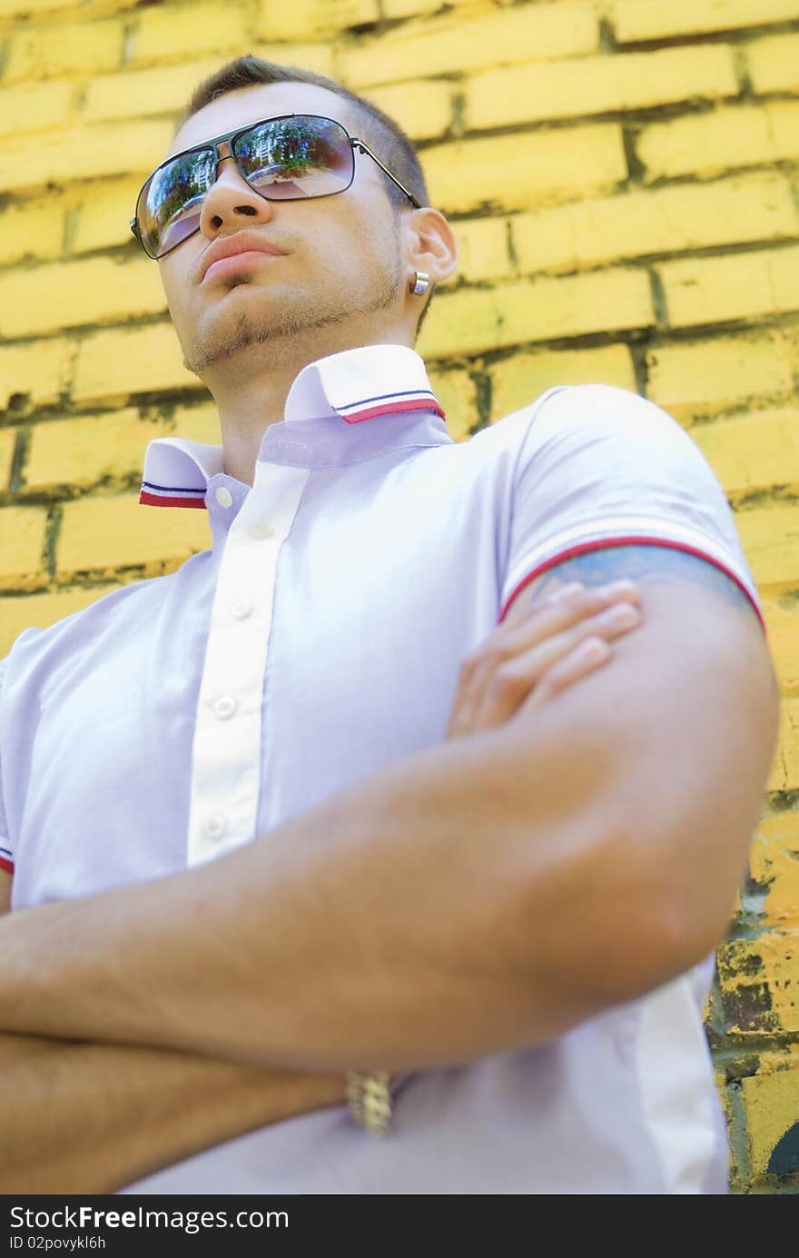 Man with big and muscled arms stands near the yellow brick wall. He wears sun glasses and earrings. Under the shirt you can see part of his tattoo. Man with big and muscled arms stands near the yellow brick wall. He wears sun glasses and earrings. Under the shirt you can see part of his tattoo.