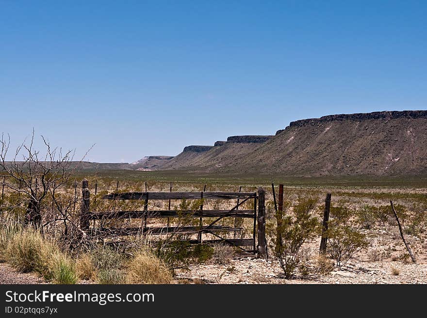 Old wooden gate in desert with mountains in background. Clear blue sky.