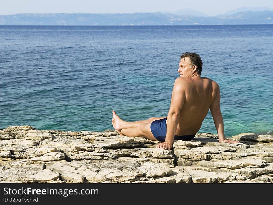 A color photo of a caucasian man sitting on rocks and relaxing next to the stunning blue turquoise sea of Greece. A color photo of a caucasian man sitting on rocks and relaxing next to the stunning blue turquoise sea of Greece.