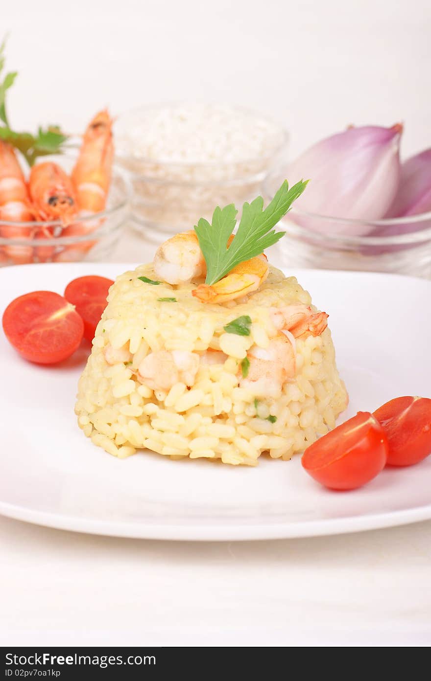 Risotto with shrimps served on a white plate and decorated with cherry tomatoes. Rice, shrimps and shallots in small glass bowls are out of focus in the background. Studio shot. Shallow DOF