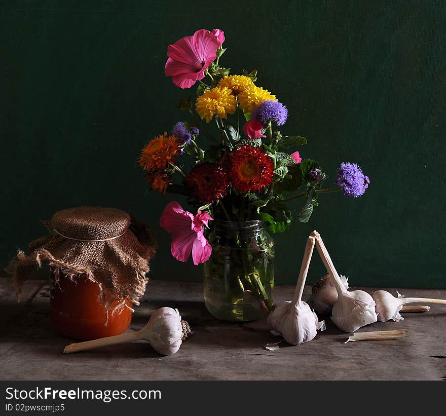 On an old table a jar stands with flowers, by the row of bank with a jam and a garlic lies. On an old table a jar stands with flowers, by the row of bank with a jam and a garlic lies