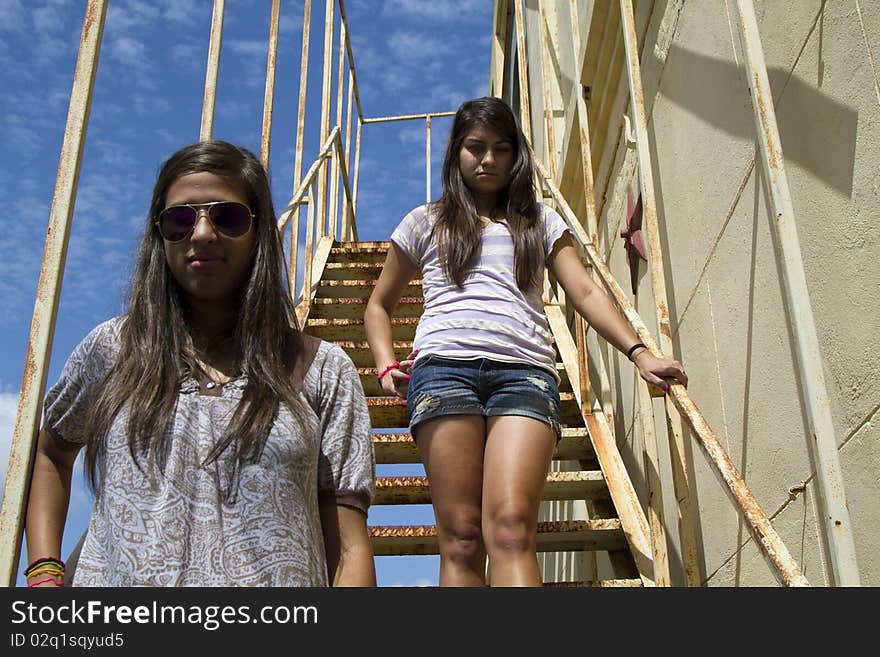 A couple of teenagers, one in sunglasses standing on a metal staircase with a bright blue sky overheard. A couple of teenagers, one in sunglasses standing on a metal staircase with a bright blue sky overheard.