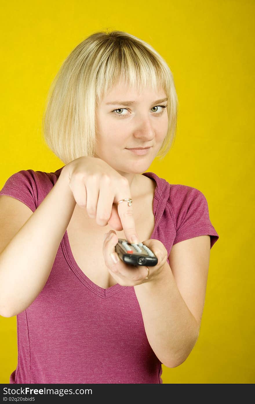 Young woman on yellow background holding a remote control designed into the frame. Finger on the other hand presses the button. Young woman on yellow background holding a remote control designed into the frame. Finger on the other hand presses the button.