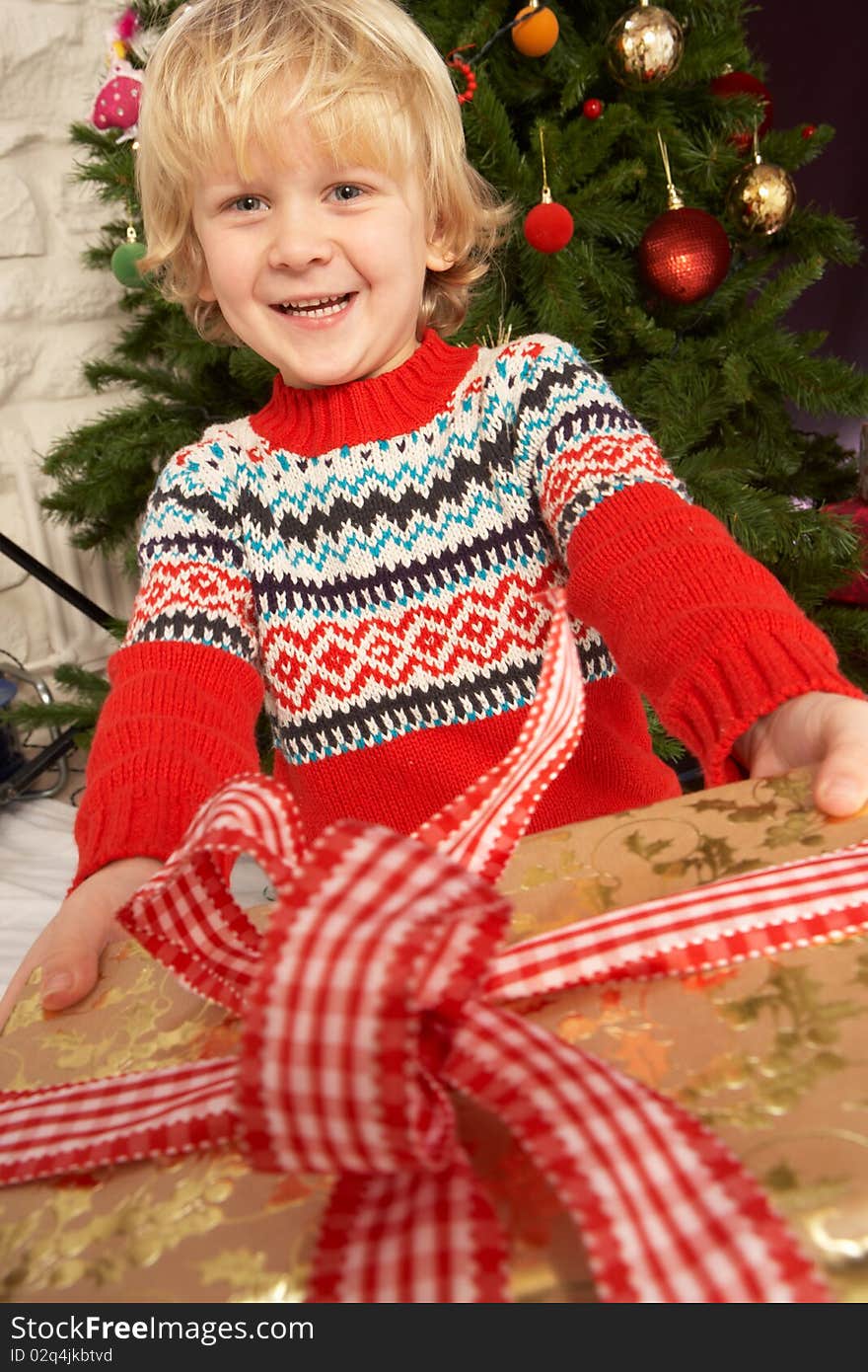 Young Boy Holding Gift In Front Of Christmas Tree Smiling