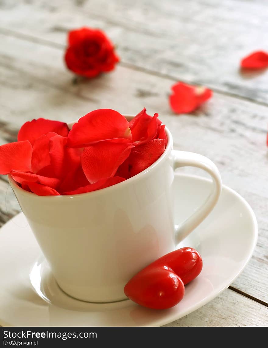 Cup of red rose petals and red heart on old wooden board