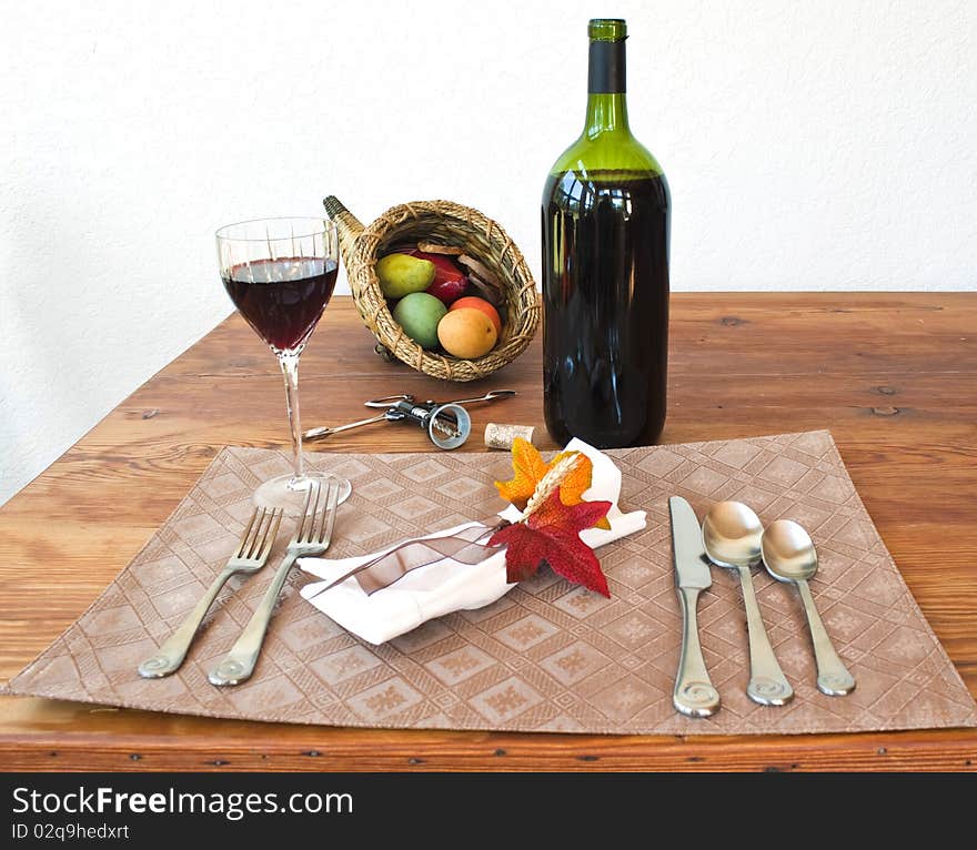 A bottle of wine, a glass of wine on a table with napkin, napkin ring with leaves, a cornucopia, silverware, placemat, and cork screw on a table. A bottle of wine, a glass of wine on a table with napkin, napkin ring with leaves, a cornucopia, silverware, placemat, and cork screw on a table.