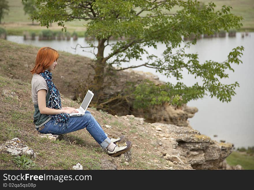 Young beautiful girl with laptop at rock near lake and tree.