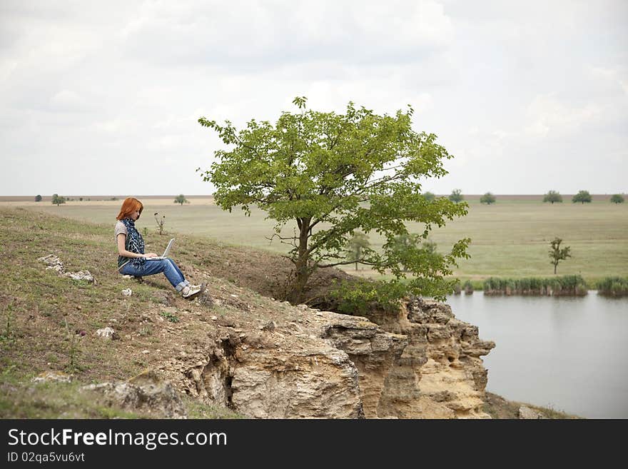 Young beautiful girl with laptop at rock near lake and tree.