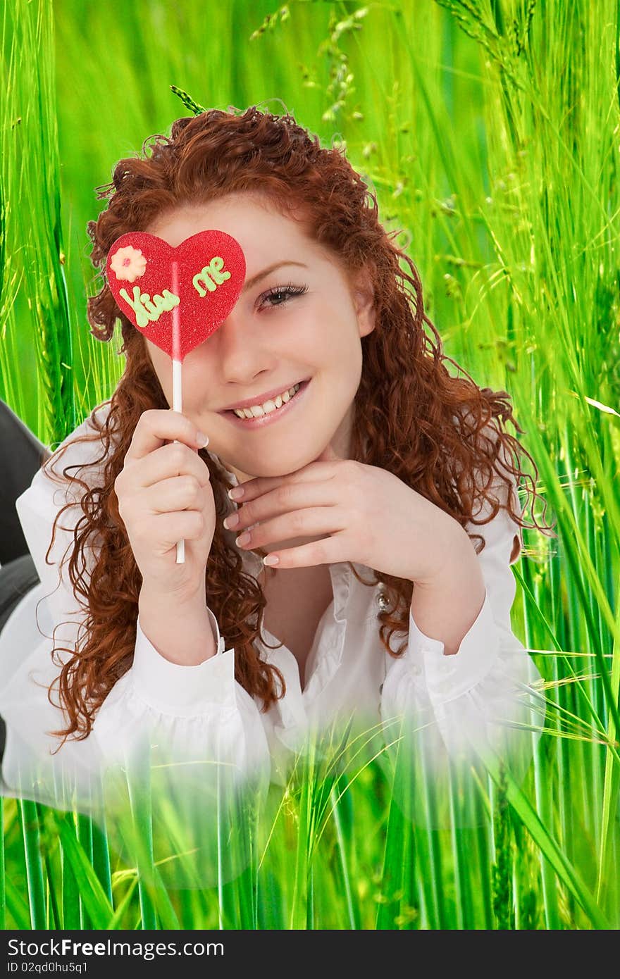 Good looking positive young caucasian girl with heartlike candy lying in green grass and smilng. Good looking positive young caucasian girl with heartlike candy lying in green grass and smilng